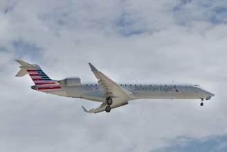 A commercial aircraft with American Eagle branding is flying against a backdrop of cloudy sky. The plane's body is mostly white with red and blue tail design. The landing gear is down, indicating it might be approaching for landing.