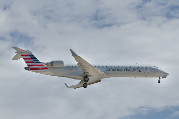 A commercial aircraft with American Eagle branding is flying against a backdrop of cloudy sky. The plane's body is mostly white with red and blue tail design. The landing gear is down, indicating it might be approaching for landing.