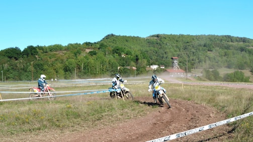 Three motorcyclists wearing protective gear are racing on a dirt track surrounded by lush green hills. A building with a red roof and a watchtower is visible in the background. The track is enclosed with tape, and the motorcyclists kick up dust as they speed around a bend.