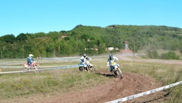 Three motorcyclists wearing protective gear are racing on a dirt track surrounded by lush green hills. A building with a red roof and a watchtower is visible in the background. The track is enclosed with tape, and the motorcyclists kick up dust as they speed around a bend.