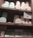 A set of wooden shelves displays various tins and boxes of champagne truffles and chocolate tiles. The truffles are stored in round tins of pastel colors, including mint green and light pink. Below, rectangular boxes labeled as chocolate tiles are also neatly arranged. A small sign is present, offering details about a 'Sweetness Rewards' program.