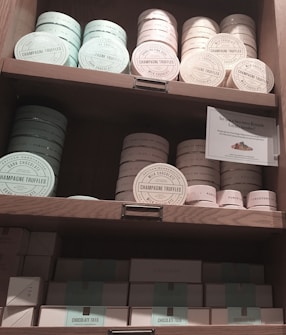 A set of wooden shelves displays various tins and boxes of champagne truffles and chocolate tiles. The truffles are stored in round tins of pastel colors, including mint green and light pink. Below, rectangular boxes labeled as chocolate tiles are also neatly arranged. A small sign is present, offering details about a 'Sweetness Rewards' program.