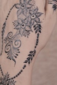 Intricate black floral henna design featuring detailed flowers, swirls, dots, and leaves on the skin. The artwork appears delicate and decorative, covering a portion of the hand with elegant patterns.