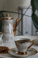 A porcelain teapot and teacup set with gold accents, decorated with floral patterns, are placed on a white surface. There are also some cookies on a plate next to the teacup. The background includes some blurred green plant leaves and a soft-focus light grey object.