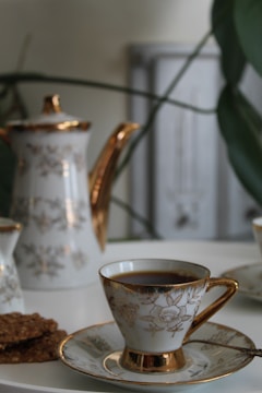 A porcelain teapot and teacup set with gold accents, decorated with floral patterns, are placed on a white surface. There are also some cookies on a plate next to the teacup. The background includes some blurred green plant leaves and a soft-focus light grey object.