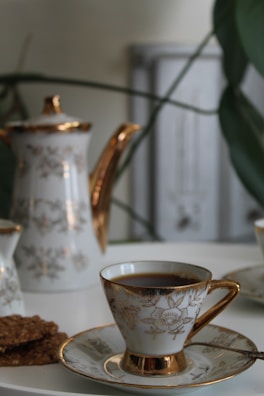 A porcelain teapot and teacup set with gold accents, decorated with floral patterns, are placed on a white surface. There are also some cookies on a plate next to the teacup. The background includes some blurred green plant leaves and a soft-focus light grey object.