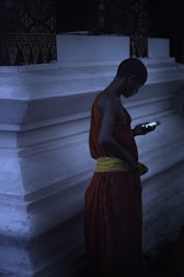 A person wearing traditional orange and yellow robes is standing in front of a white concrete structure, looking at a smartphone in their hand. The setting appears to be dimly lit, creating a serene and contemplative atmosphere.