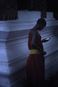 A person wearing traditional orange and yellow robes is standing in front of a white concrete structure, looking at a smartphone in their hand. The setting appears to be dimly lit, creating a serene and contemplative atmosphere.