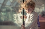 A young child with curly hair is looking intently at their reflection in a glass surface. The child is wearing a light-colored hoodie. The background appears to be a large indoor space with natural light streaming in, possibly an atrium or a terminal.