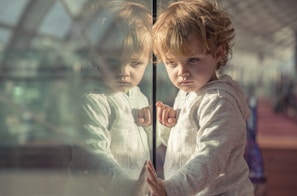 A young child with curly hair is looking intently at their reflection in a glass surface. The child is wearing a light-colored hoodie. The background appears to be a large indoor space with natural light streaming in, possibly an atrium or a terminal.