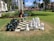 A life-sized outdoor chess set is arranged on a checkered board laid on a grassy area. A person is bent over, interacting with one of the large chess pieces. The scene is set in a sunlit park with palm trees and nearby buildings visible in the background. A sign next to the chess set reads 'Game Time'.