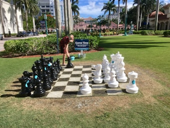 A life-sized outdoor chess set is arranged on a checkered board laid on a grassy area. A person is bent over, interacting with one of the large chess pieces. The scene is set in a sunlit park with palm trees and nearby buildings visible in the background. A sign next to the chess set reads 'Game Time'.