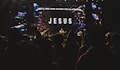 A large, dimly lit concert setting with a crowd of people watching performers on stage. The backdrop features a large screen displaying the word 'JESUS' and a message about singing for eternity. Raised hands in the audience suggest engagement and enthusiasm.