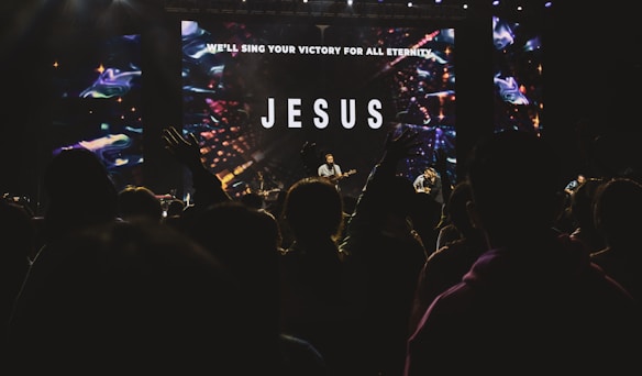 A large, dimly lit concert setting with a crowd of people watching performers on stage. The backdrop features a large screen displaying the word 'JESUS' and a message about singing for eternity. Raised hands in the audience suggest engagement and enthusiasm.