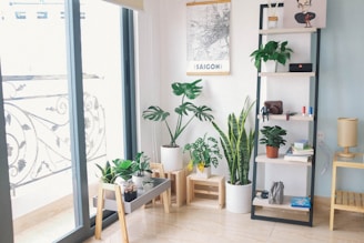 A cozy indoor space features a collection of potted plants including monstera and snake plants, set near a glass door leading to a balcony with ornate metal railings. A minimalist shelving unit displays small items such as books, decorative pieces, and a compact radio. The wall showcases a map poster labeled 'SAIGON,' adding a personal touch to the room. A small wooden table supports more plants, while a light-colored floor lamp adds warmth to the corner.