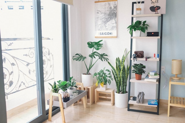 A cozy indoor space features a collection of potted plants including monstera and snake plants, set near a glass door leading to a balcony with ornate metal railings. A minimalist shelving unit displays small items such as books, decorative pieces, and a compact radio. The wall showcases a map poster labeled 'SAIGON,' adding a personal touch to the room. A small wooden table supports more plants, while a light-colored floor lamp adds warmth to the corner.