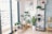 A cozy indoor space features a collection of potted plants including monstera and snake plants, set near a glass door leading to a balcony with ornate metal railings. A minimalist shelving unit displays small items such as books, decorative pieces, and a compact radio. The wall showcases a map poster labeled 'SAIGON,' adding a personal touch to the room. A small wooden table supports more plants, while a light-colored floor lamp adds warmth to the corner.
