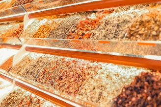 A vibrant display of various dried herbs and spices arranged in transparent containers on angled shelves. The collection includes an assortment of colors and textures from different varieties of dried produce.