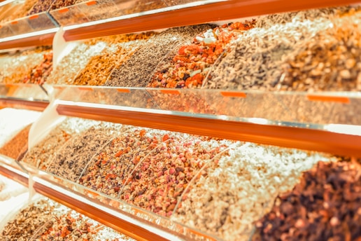 A vibrant display of various dried herbs and spices arranged in transparent containers on angled shelves. The collection includes an assortment of colors and textures from different varieties of dried produce.