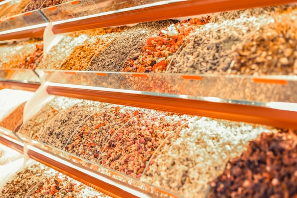 A vibrant display of various dried herbs and spices arranged in transparent containers on angled shelves. The collection includes an assortment of colors and textures from different varieties of dried produce.