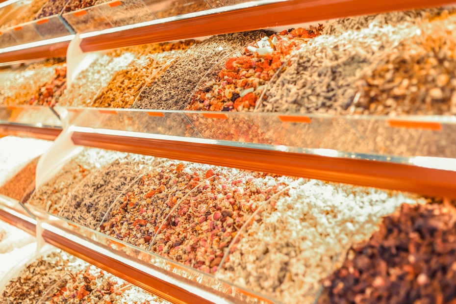 A vibrant display of various dried herbs and spices arranged in transparent containers on angled shelves. The collection includes an assortment of colors and textures from different varieties of dried produce.