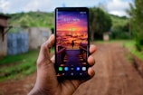 A smartphone is being held in a hand with a vibrant, colorful wallpaper depicting a sunset over a city with birds in the sky and a wooden walkway leading to a railing. The background shows an outdoor setting with a dirt path and greenery, slightly blurred.
