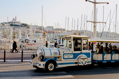 A small tourist train painted in white and blue is traveling along a waterfront area with numerous docked boats. The area is bustling with people walking nearby. In the background, a hill with a large building at the top is visible, giving a scenic view of the city skyline.