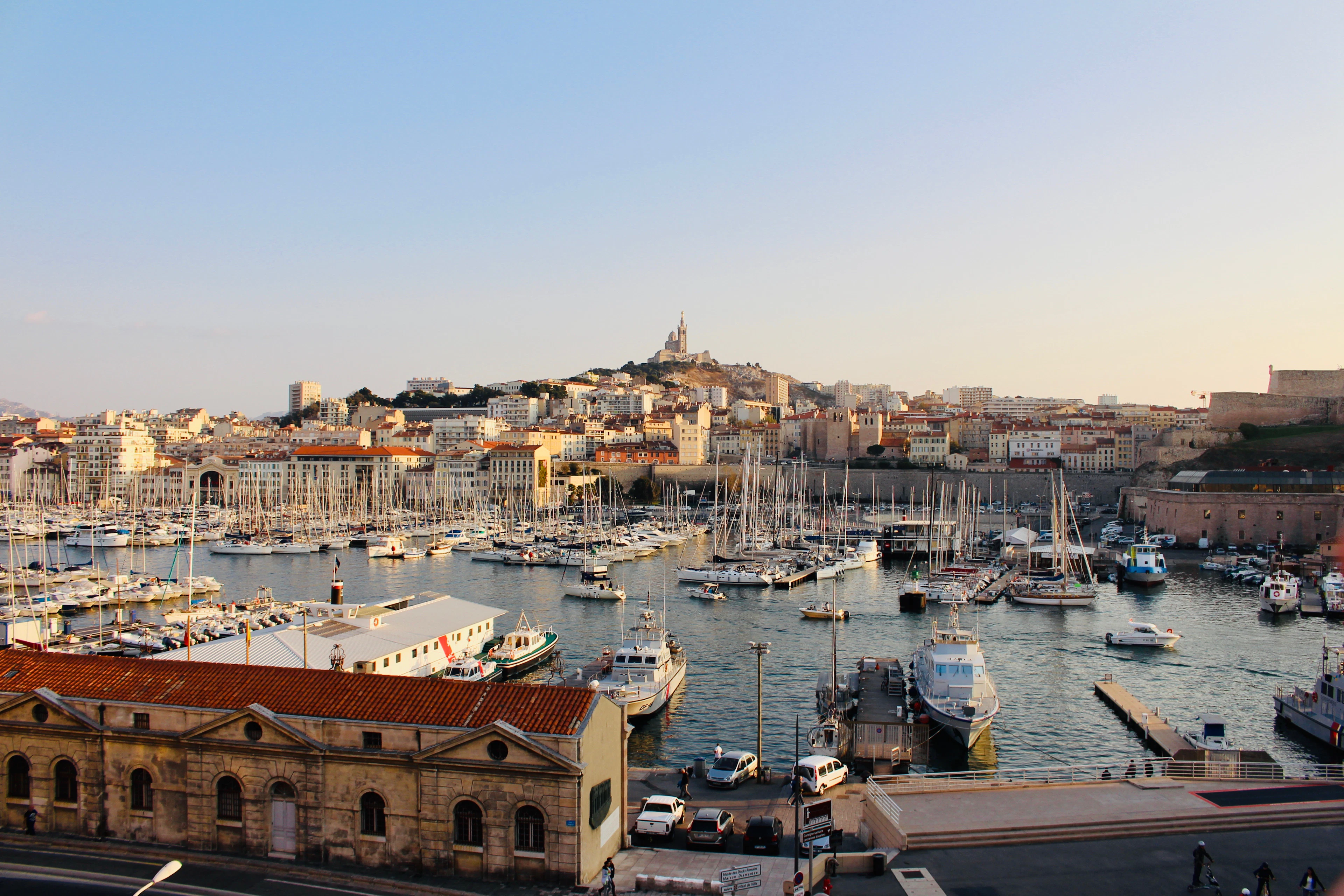 Sailboats moored in Marseille's Vieux-Port at golden hour, with the hilltop basilica of Notre-Dame de la Garde rising above the city