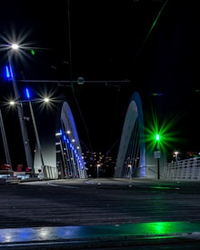 A modern bridge with a sleek design is illuminated at night, featuring bright lights in blue and green hues. The structure has large, white arches and is surrounded by a dark sky. The bridge's surface appears smooth and metallic, with visible railings and some distant city lights in the background.