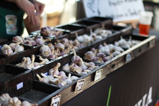 A market stall displays various types of garlic organized in small wooden compartments. Several piles of garlic bulbs, with a mix of white and purple hues, are arranged neatly. A person’s hands are visible in the background, appearing to sort or handle the garlic. A chalkboard-style sign is partially visible, possibly indicating the types or prices of the garlic.