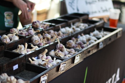 A market stall displays various types of garlic organized in small wooden compartments. Several piles of garlic bulbs, with a mix of white and purple hues, are arranged neatly. A person’s hands are visible in the background, appearing to sort or handle the garlic. A chalkboard-style sign is partially visible, possibly indicating the types or prices of the garlic.