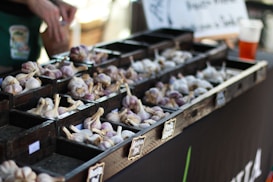 A market stall displays various types of garlic organized in small wooden compartments. Several piles of garlic bulbs, with a mix of white and purple hues, are arranged neatly. A person&rsquo;s hands are visible in the background, appearing to sort or handle the garlic. A chalkboard-style sign is partially visible, possibly indicating the types or prices of the garlic.