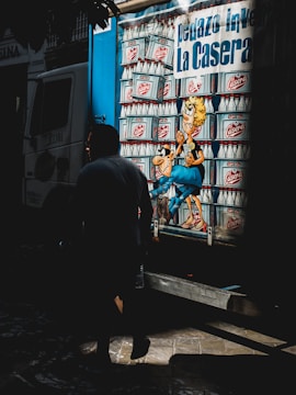 A truck with vibrant cartoon-like artwork featuring a stack of glass bottles. A cartoon character with exaggerated features is prominently displayed, surrounded by these bottles. A person is walking past in the foreground, casting a shadow.