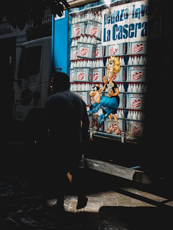 A truck with vibrant cartoon-like artwork featuring a stack of glass bottles. A cartoon character with exaggerated features is prominently displayed, surrounded by these bottles. A person is walking past in the foreground, casting a shadow.