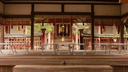 A traditional Japanese shrine features wooden architecture, intricate railings, and hanging golden ornaments. The interior appears serene, with symmetrical structures and natural light softly illuminating the space. Greenery is visible outside, providing a tranquil backdrop.