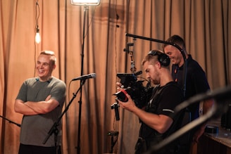 Three people are gathered in a room, with one man smiling and standing near a microphone. Another person is operating a professional video camera, wearing large headphones. A third person appears in the background, looking towards the camera operator. The setting has warm lighting and drawn curtains in the background.