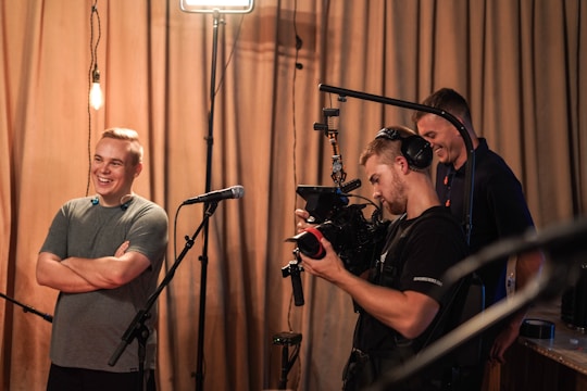 Three people are gathered in a room, with one man smiling and standing near a microphone. Another person is operating a professional video camera, wearing large headphones. A third person appears in the background, looking towards the camera operator. The setting has warm lighting and drawn curtains in the background.