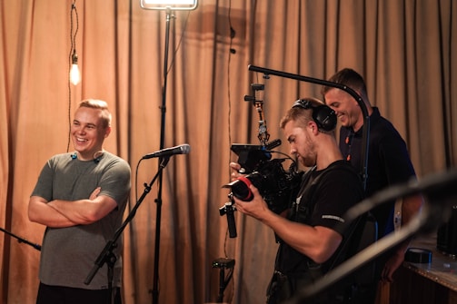Three people are gathered in a room, with one man smiling and standing near a microphone. Another person is operating a professional video camera, wearing large headphones. A third person appears in the background, looking towards the camera operator. The setting has warm lighting and drawn curtains in the background.