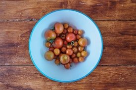 A collection of various sized ripe tomatoes is placed in a light blue bowl. The bowl rests on a rustic wooden surface, creating a contrast between the vibrant red and orange hues of the tomatoes and the subdued blue of the bowl. The wooden texture adds a natural and warm backdrop.