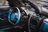 A luxurious car interior featuring a high-end steering wheel with blue and black leather accents. The dashboard presents a sophisticated design with multiple analog dials, a large digital display, and various control buttons. Carbon fiber elements are prominent, enhancing the sleek and modern aesthetic.