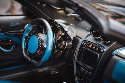 A luxurious car interior featuring a high-end steering wheel with blue and black leather accents. The dashboard presents a sophisticated design with multiple analog dials, a large digital display, and various control buttons. Carbon fiber elements are prominent, enhancing the sleek and modern aesthetic.