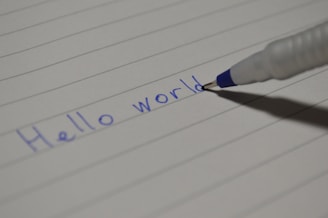 A ballpoint pen is writing the phrase 'Hello world' on a sheet of lined paper. The ink is blue, and the paper is white with horizontal lines.