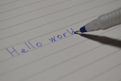 A ballpoint pen is writing the phrase 'Hello world' on a sheet of lined paper. The ink is blue, and the paper is white with horizontal lines.