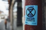 A blue sticker with a black hourglass symbol and the text 'THERE IS NO PLANET B' is affixed to a red pole. The background features a blurred street scene, suggesting an urban environment.