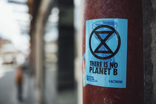 A blue sticker with a black hourglass symbol and the text 'THERE IS NO PLANET B' is affixed to a red pole. The background features a blurred street scene, suggesting an urban environment.