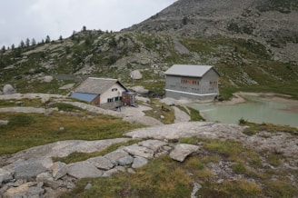 Two buildings are situated in a mountainous landscape with patches of green grass and rocky terrain. A small lake with pale turquoise water is visible next to the buildings, and a few trees are scattered on the hill. The sky is overcast, adding a grayish tint to the scene.