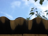 A close-up view of a wavy metal roof edge against a bright blue sky. In the background, blurred green leaves partially visible, adding a natural contrast.