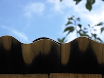 A close-up view of a wavy metal roof edge against a bright blue sky. In the background, blurred green leaves partially visible, adding a natural contrast.