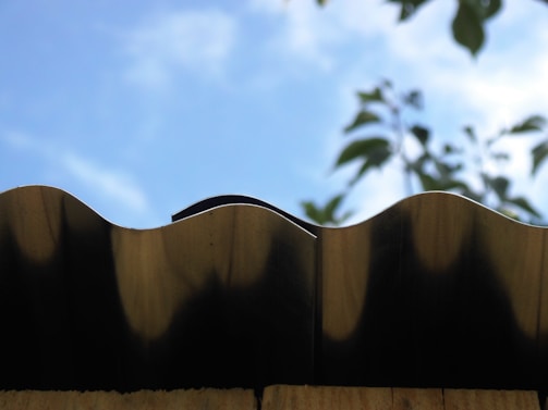 A close-up view of a wavy metal roof edge against a bright blue sky. In the background, blurred green leaves partially visible, adding a natural contrast.