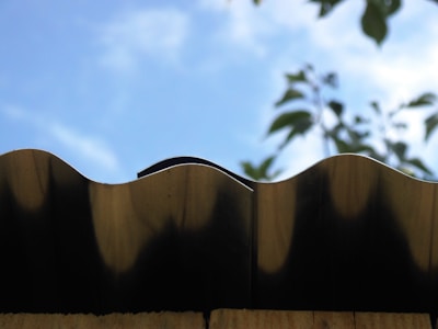 A close-up view of a wavy metal roof edge against a bright blue sky. In the background, blurred green leaves partially visible, adding a natural contrast.